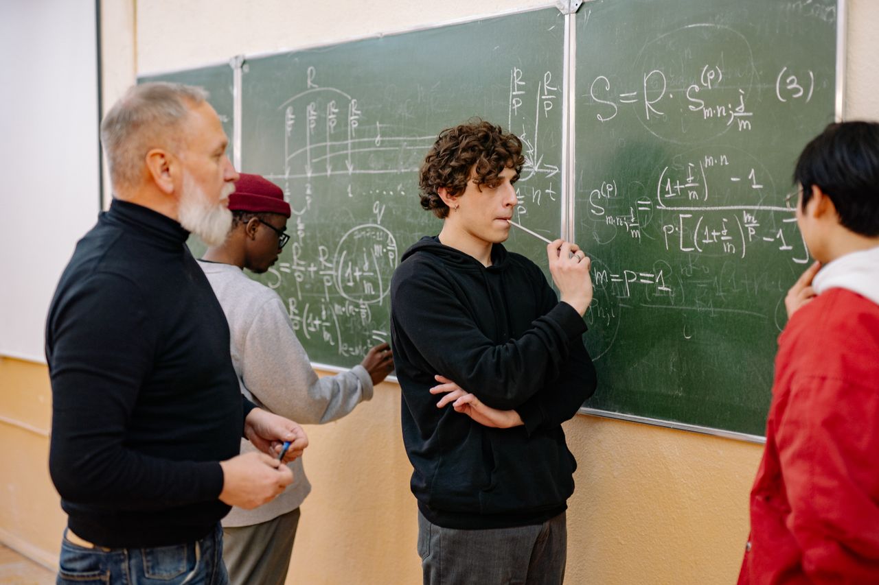 our-stories Group of students and professor discussing complex equations on a chalkboard.